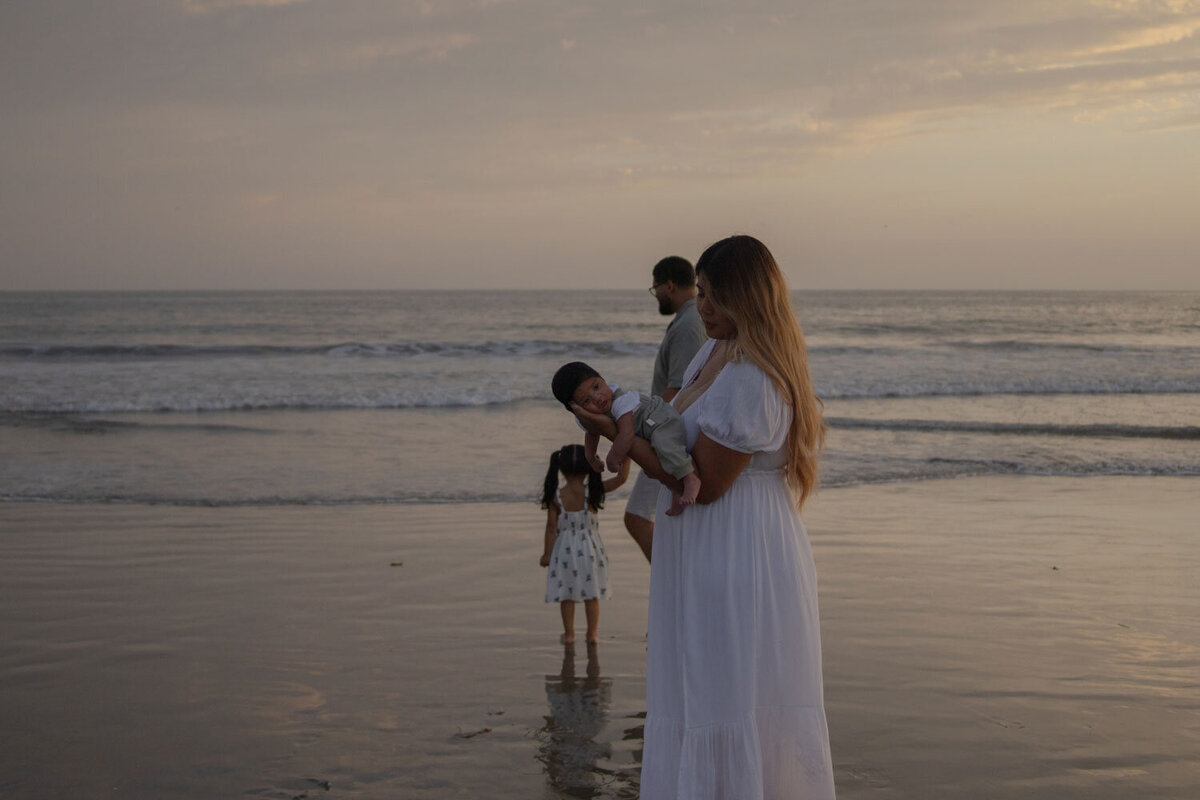 Family standing by the ocean at dusk, the mother holding the baby