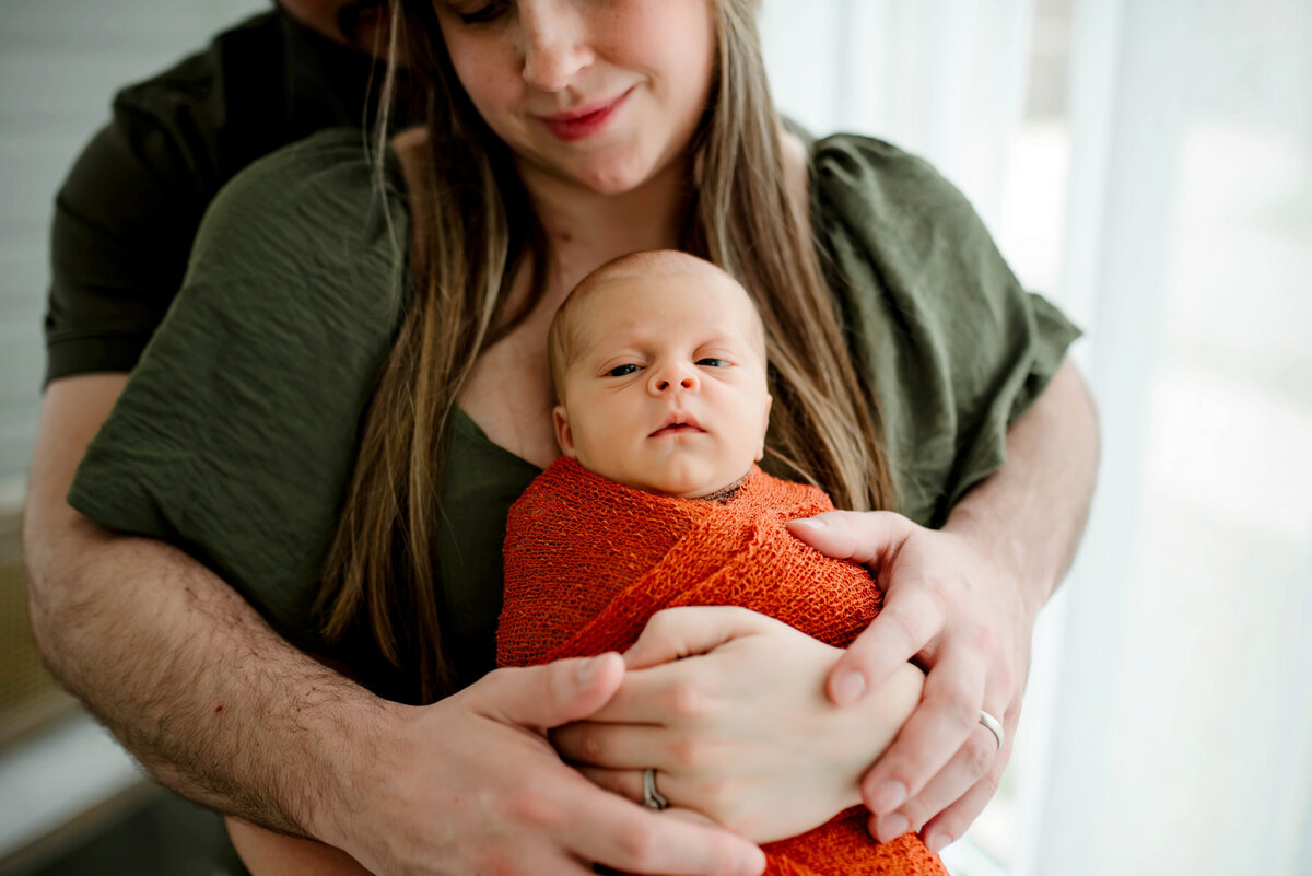mother holding his newborn baby wrapped in a soft orange blanket during an intimate in-home session in McKinney, Texas.