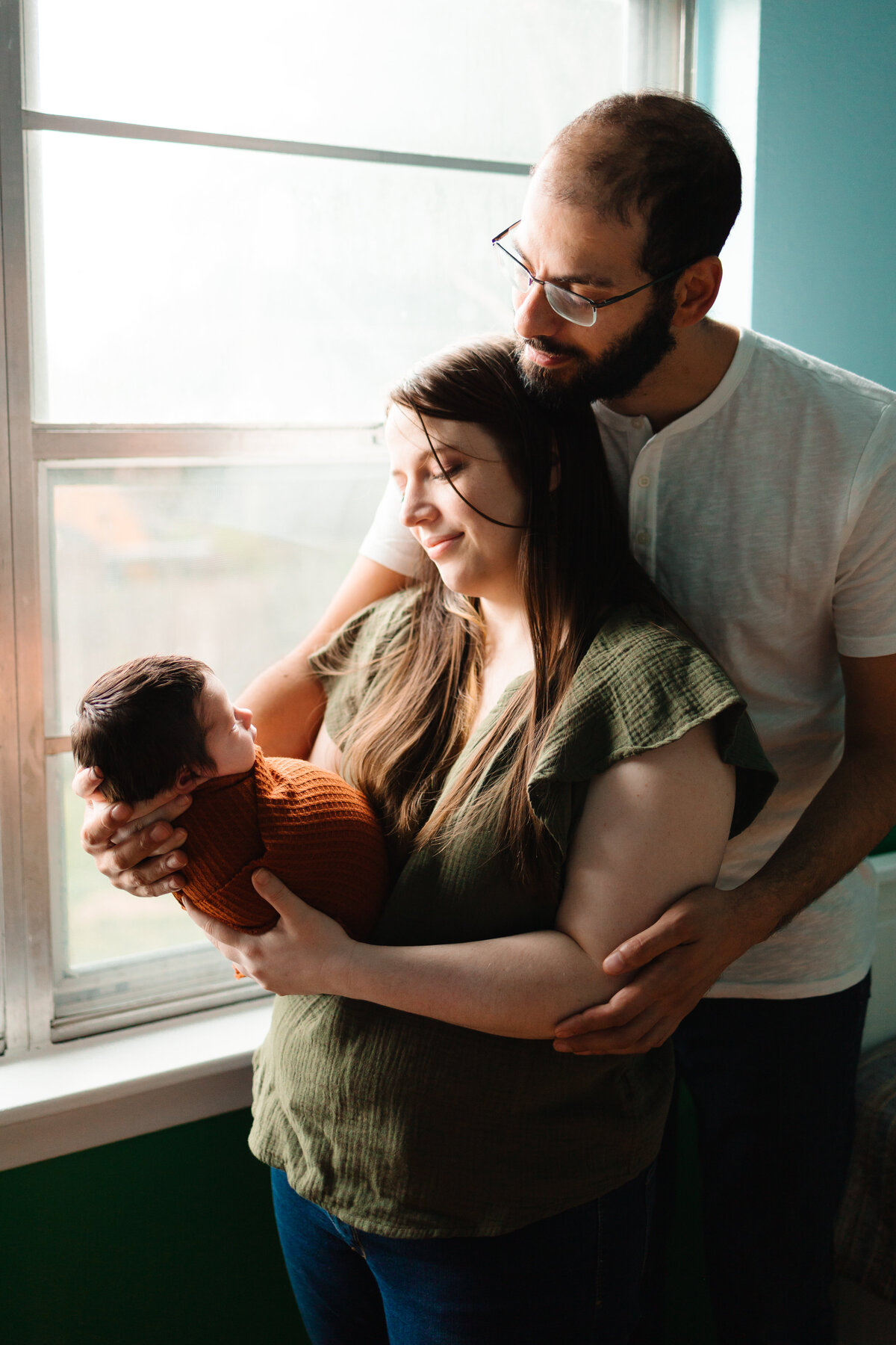 Picture of a family with a newborn baby, the parents are holding him in their arms with a dark orange blanket. The mother is dressed in a green blouse and jeans, and the father in a white one.