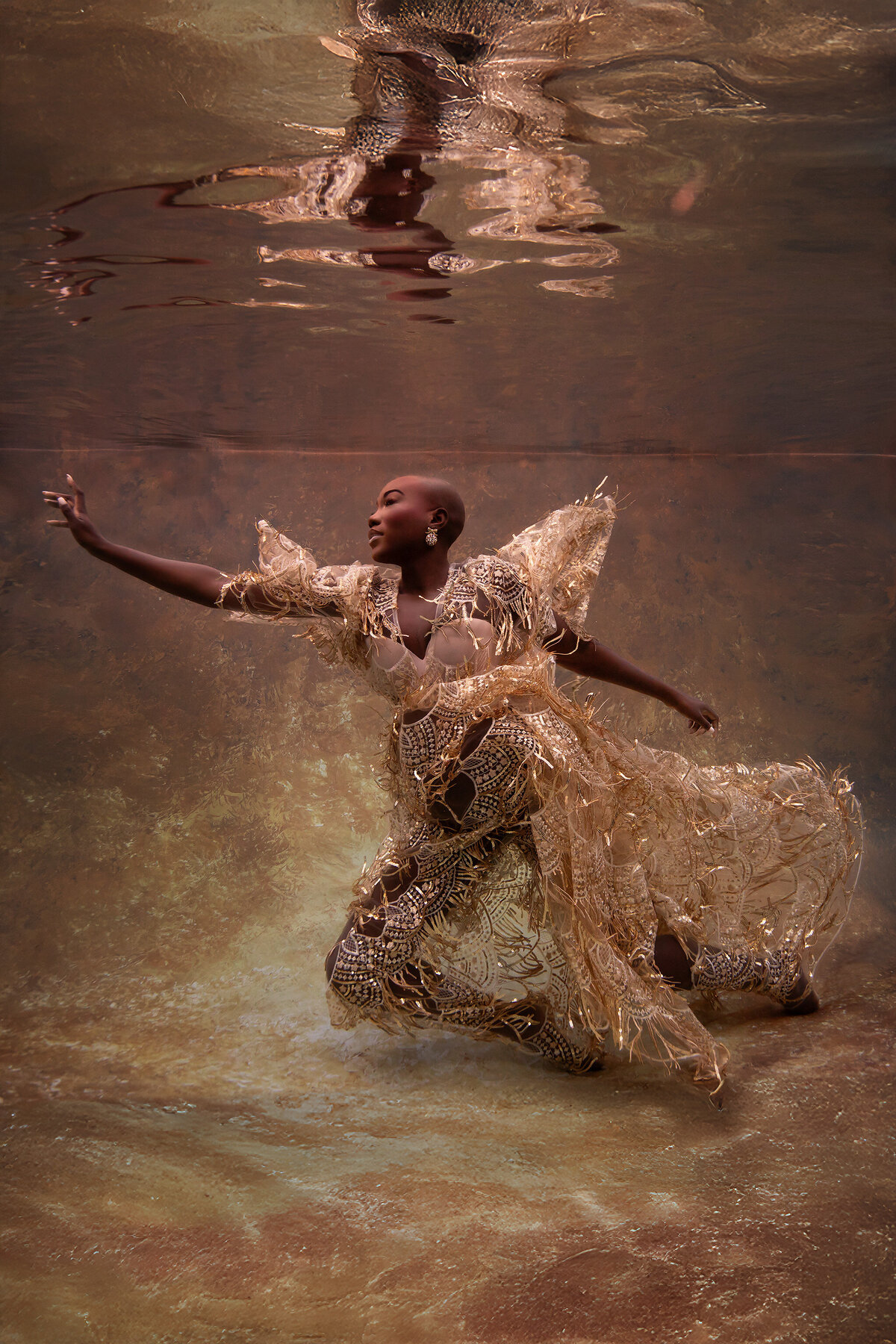 A woman dressed in an opulent gold-fringed lace gown gracefully poses underwater against a glowing golden backdrop. Her outstretched arm and elegant posture convey power and elegance. The intricate detailing of her gown shimmers beautifully in the warm, ambient light, enhancing the luxurious feel of the scene.