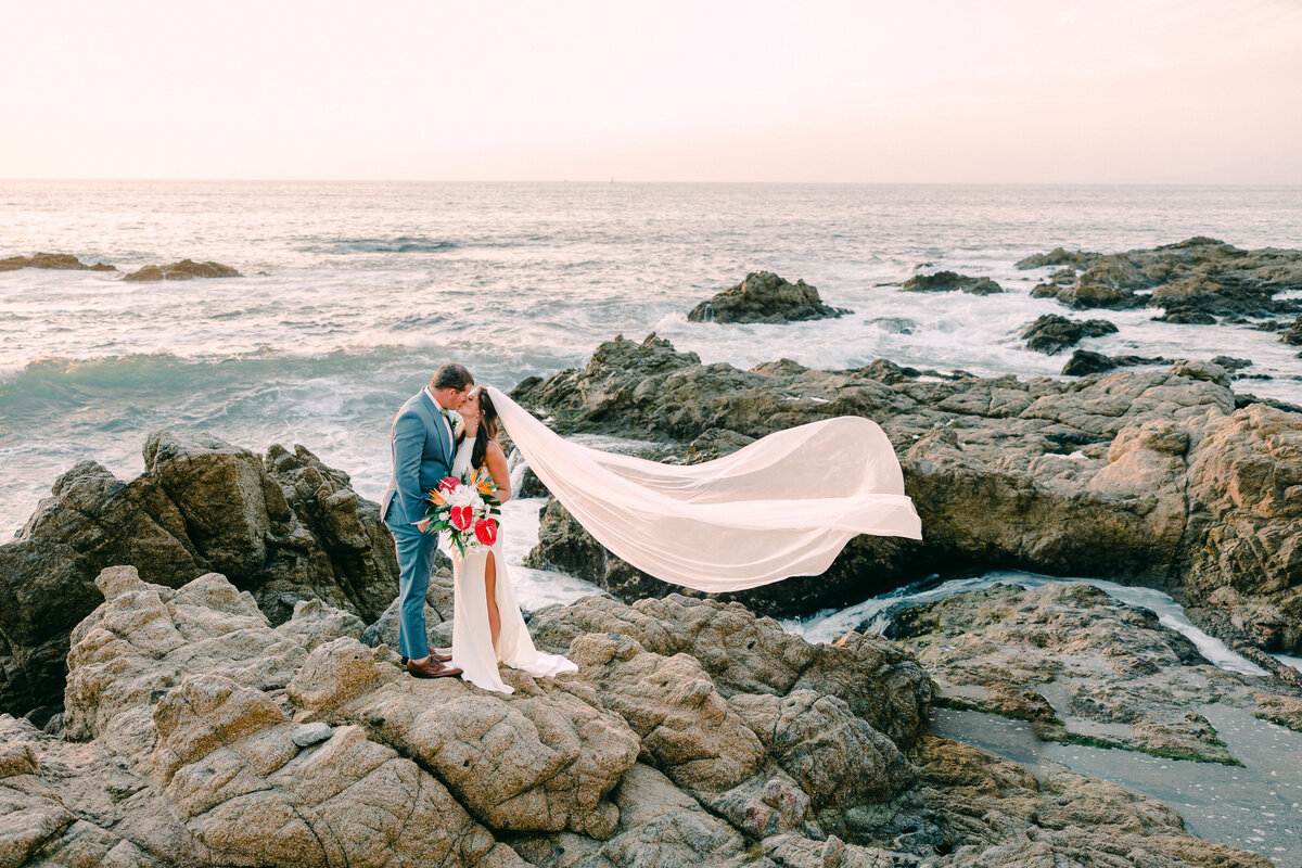Bride and groom kiss, with their veil, blowing in the wind on a rocky beach.