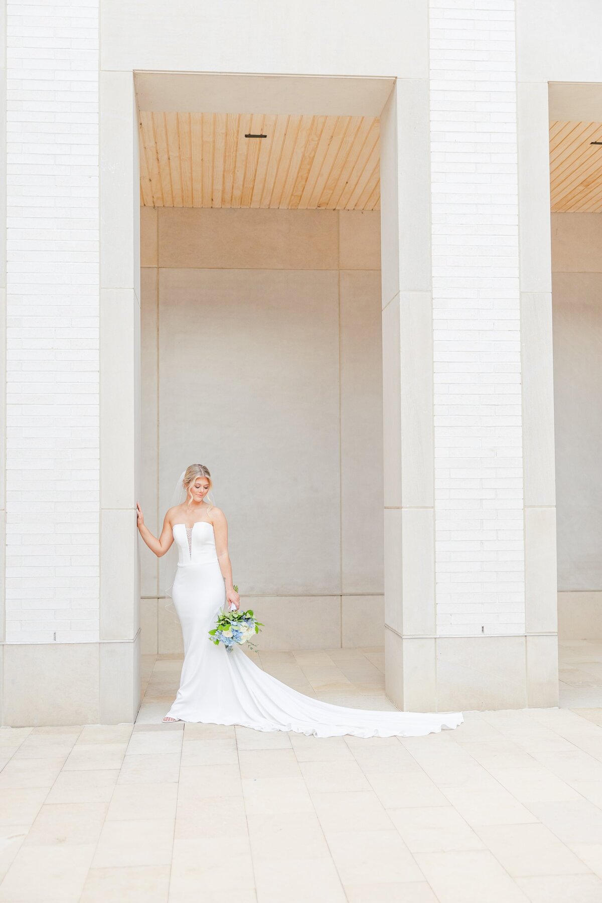 A bride smiles down her shoulder to her bouquet at her side while leaning on large columns captured by an Arkansas wedding photographer