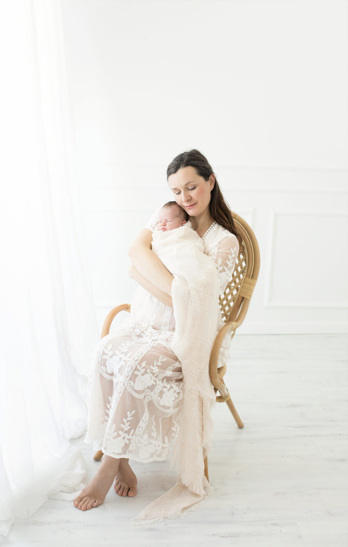 Babyshooting mit Mama und Ihrem Neugeborenen auf dem Arm vor weißer Kulisse im weißen Kleid auf einem Rattansessel