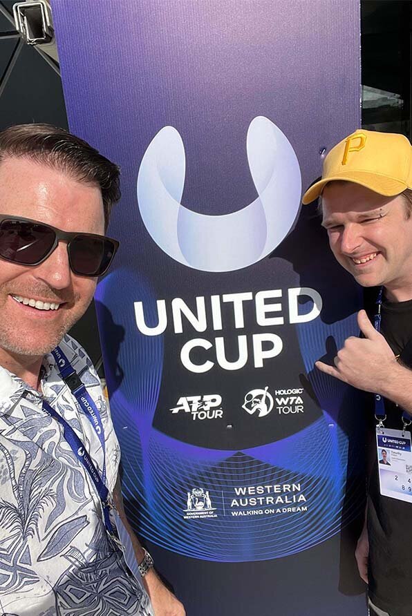 Two event attendees smiling in front of a large United Cup promotional banner featuring ATP and WTA logos