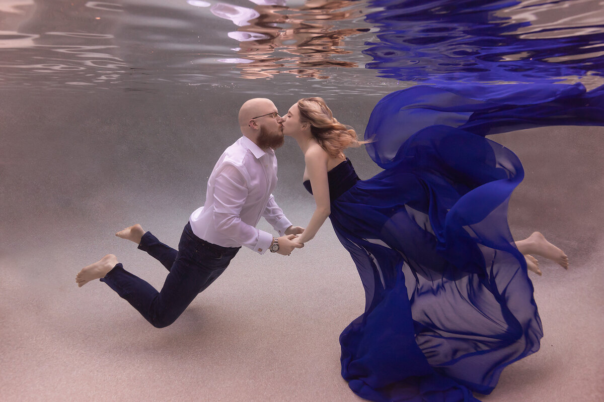 A breathtaking underwater maternity photo of a couple sharing a kiss while holding hands. The woman, wearing a flowing blue gown, floats gracefully beside her partner dressed in a white shirt and jeans. This Dallas underwater maternity photographer captures the beauty of connection and elegance in an ethereal underwater scene.