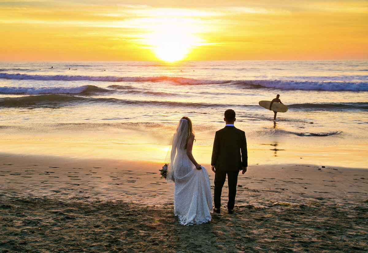 Bride and groom standing on Del Mar Beach at sunset, watching a surfer walk into the waves