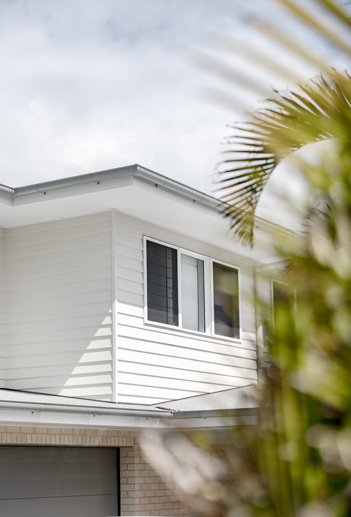 Close-up of a modern white house exterior with palm leaves partially in view, showcasing the elegance of Luxury Home Design Newcastle against a cloudy sky backdrop.