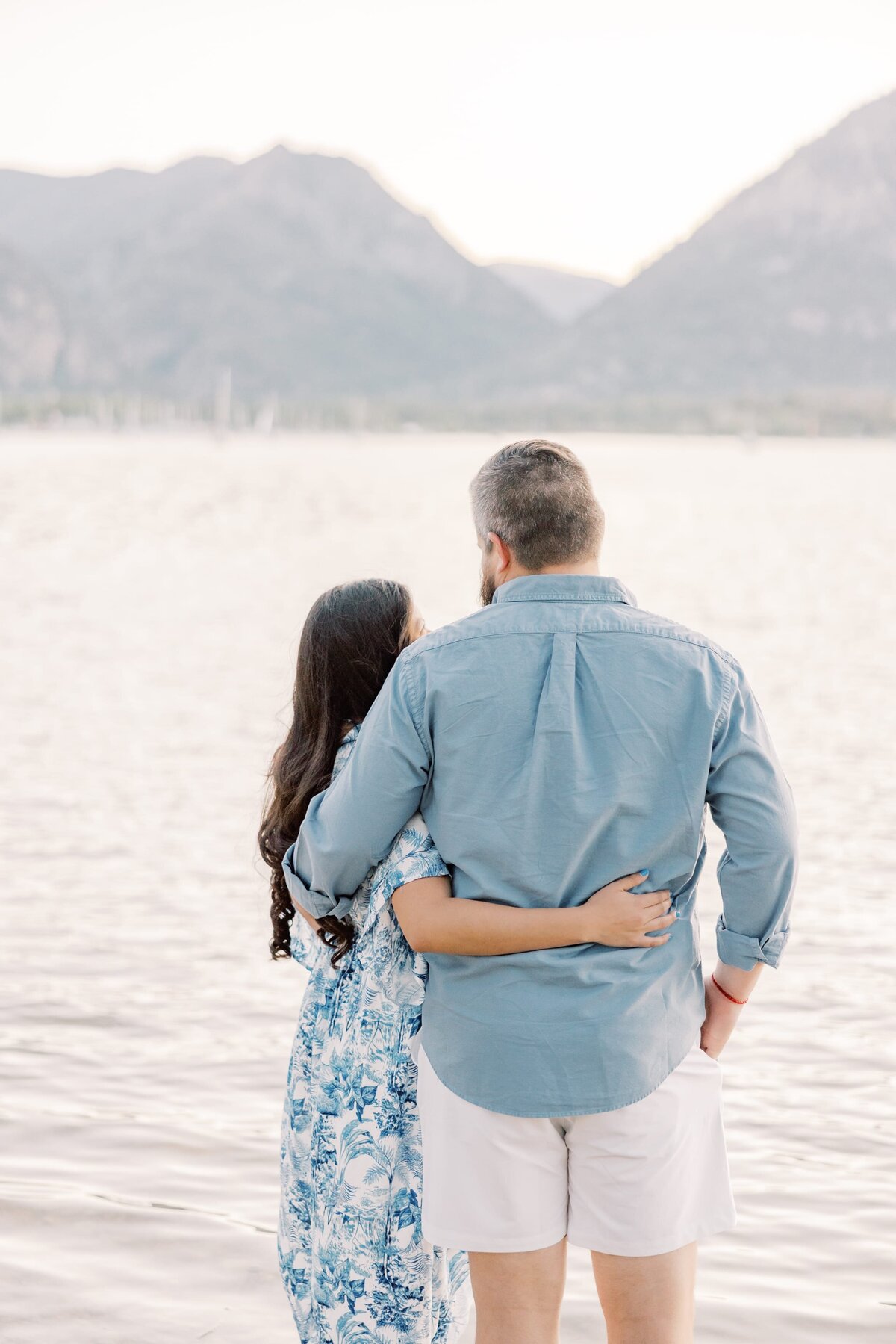 The boyfriend and girlfriend are wrapped arm in arm looking out at the scenery. They are on the shores of Lake Dillon with the mountains behind them. 