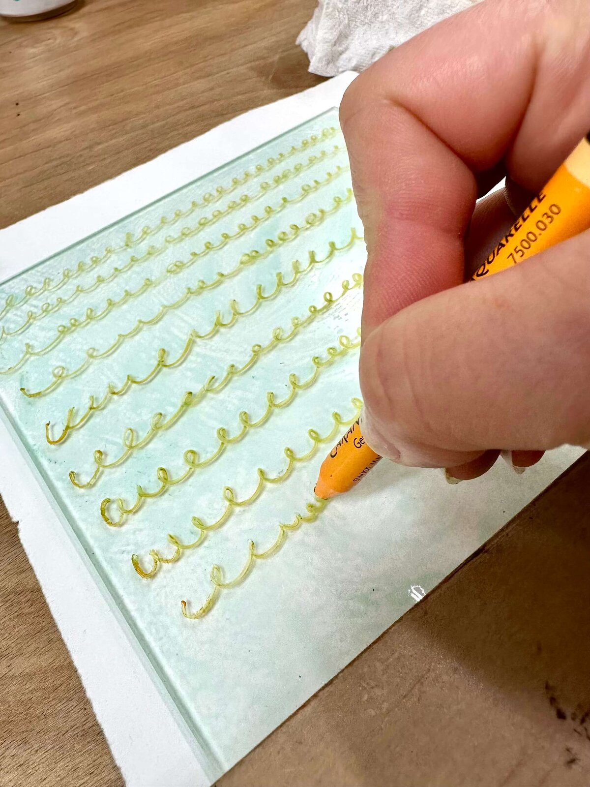 Close-up of a hand drawing yellow curly lines with a water-soluble crayon on plexiglass, showing the hands-on creativity of the monoprinting process.