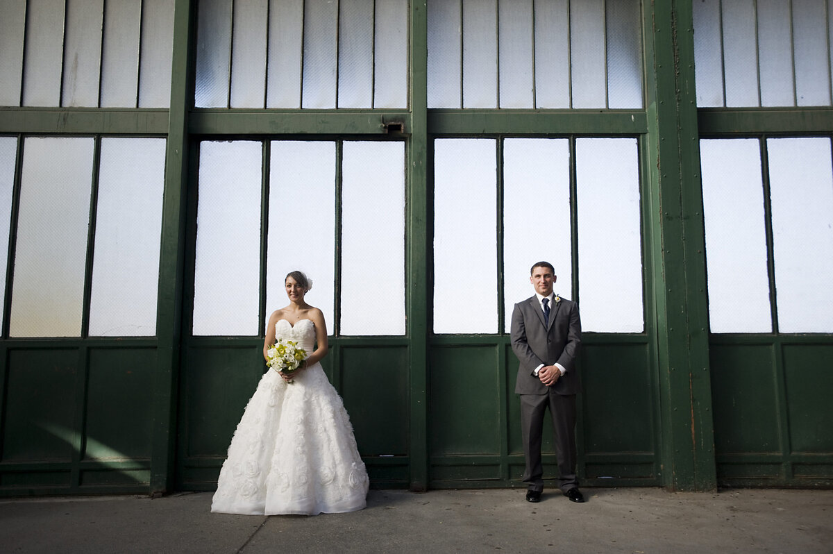 A newlywed couple standing a few feet apart in front of an industrial style building 