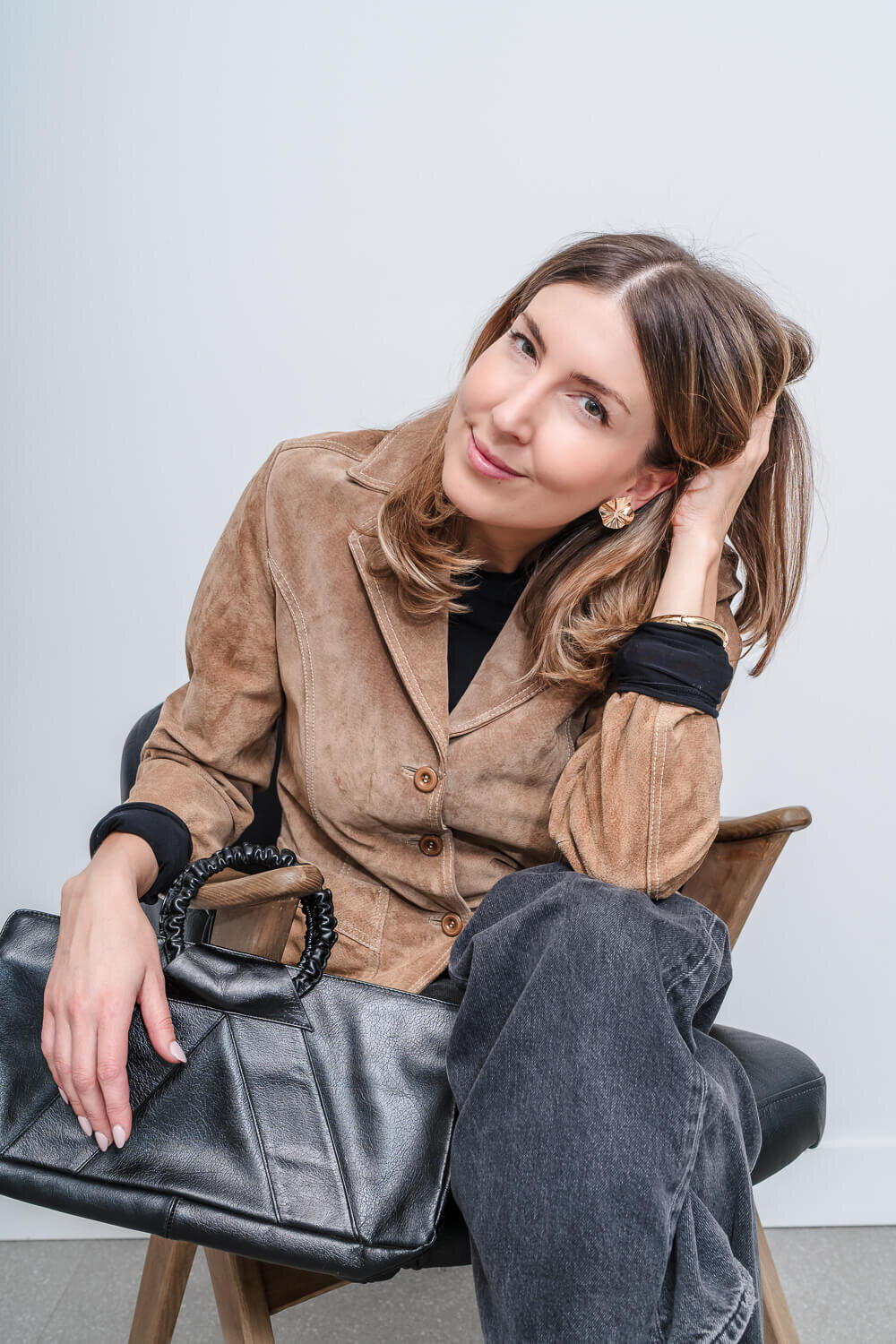 Smiling woman in tan suede jacket seated on chair holding black leather purse on her lap.