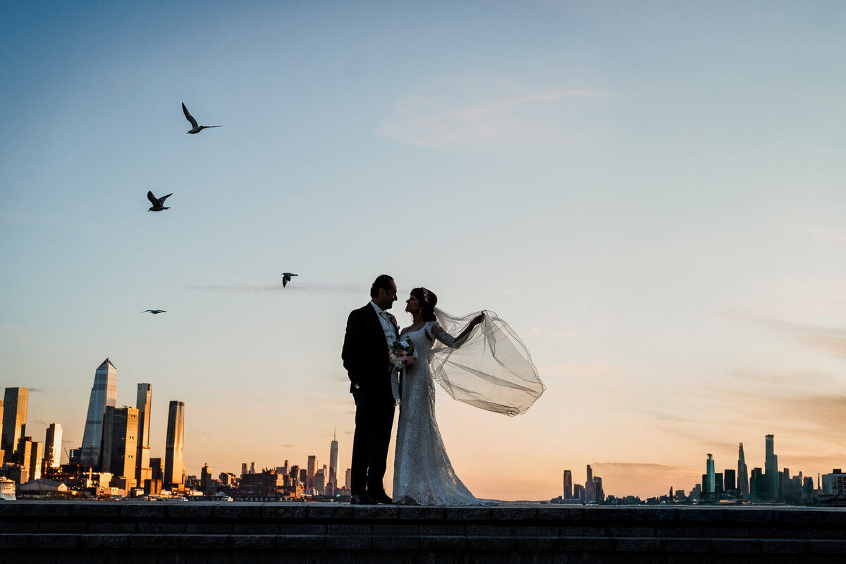 Bride and groom silhouetted against the New York City skyline at sunset as birds fly overhead and the bride’s veil catches the wind.