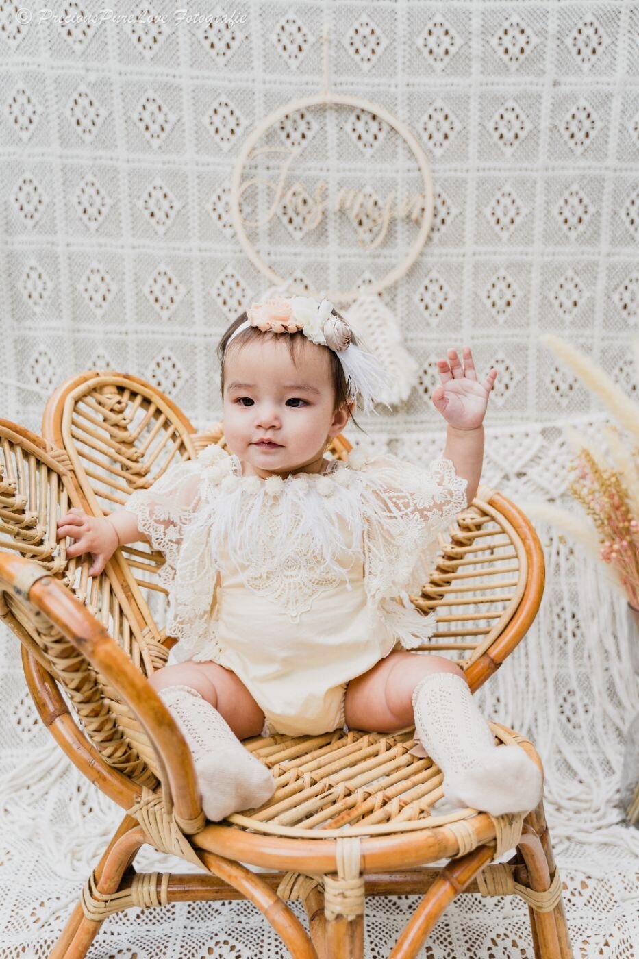 Baby girl sitting in a wicker chair, dressed in a cream lace romper and floral headband, raising one hand in a playful gesture.