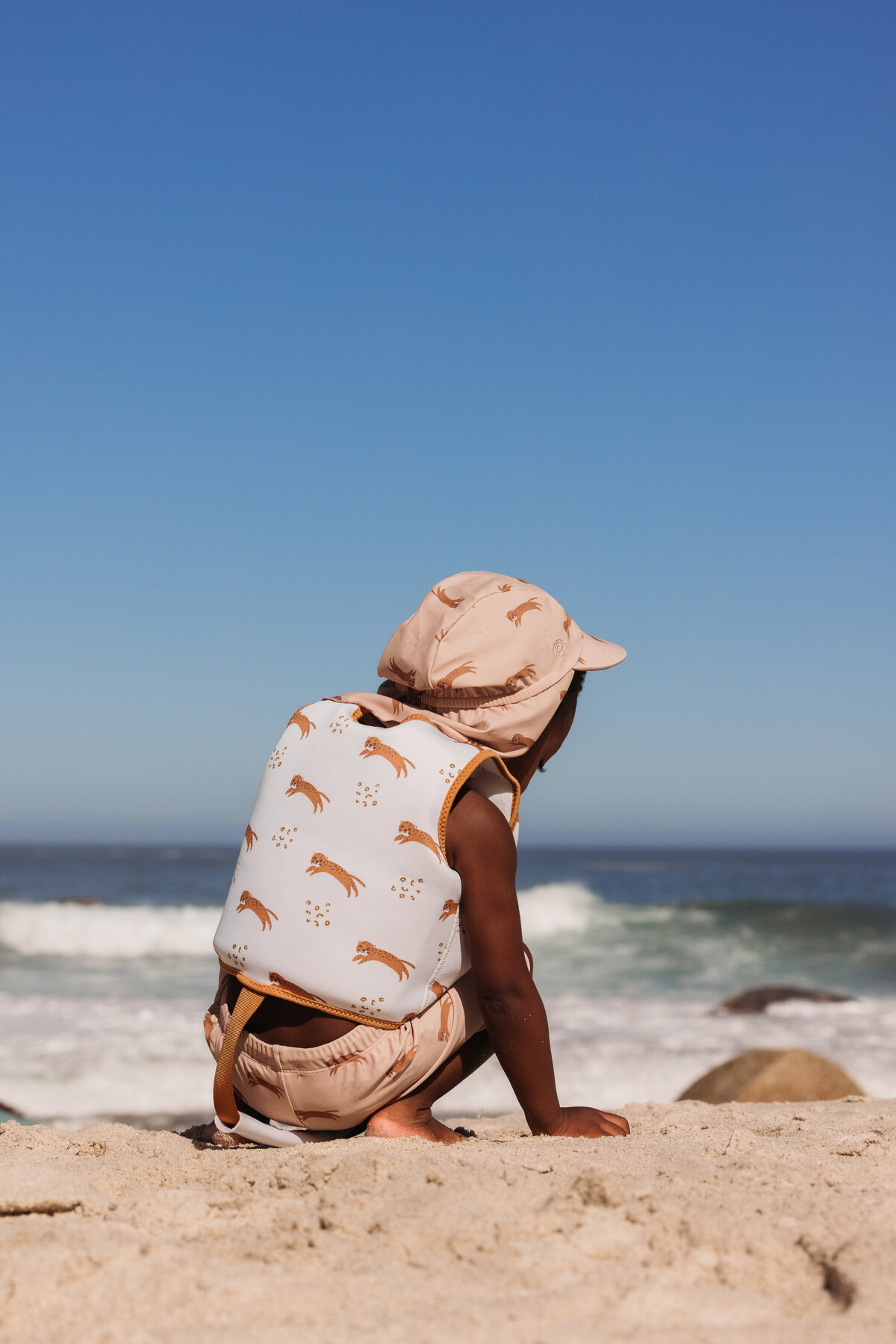 Young child wearing Swim Essentials leopard-print swim vest and sun hat, sitting on the beach while looking out at the ocean in Cape Town.