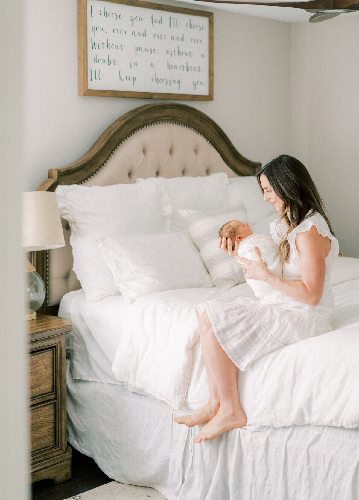 Portrait of a woman wearing a white dress sitting holding a swaddled baby in her bed room