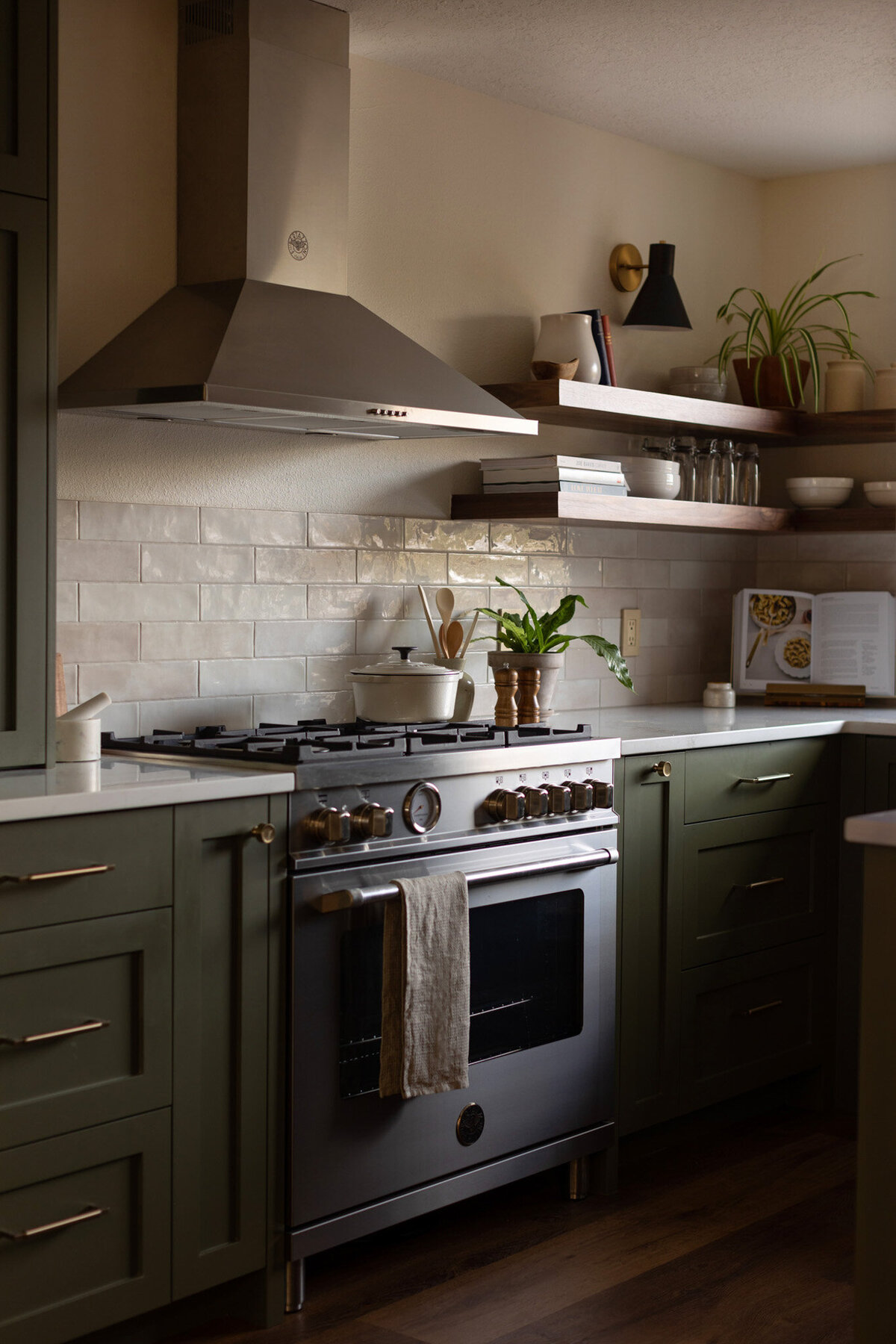 Kitchen with green cabinets, white countertops, and beige subway tile backsplash. Walnut floating shelves styled with books, plates, alcohol, and glasses. Black barstools at the island. Black pendant lights hang above the island.