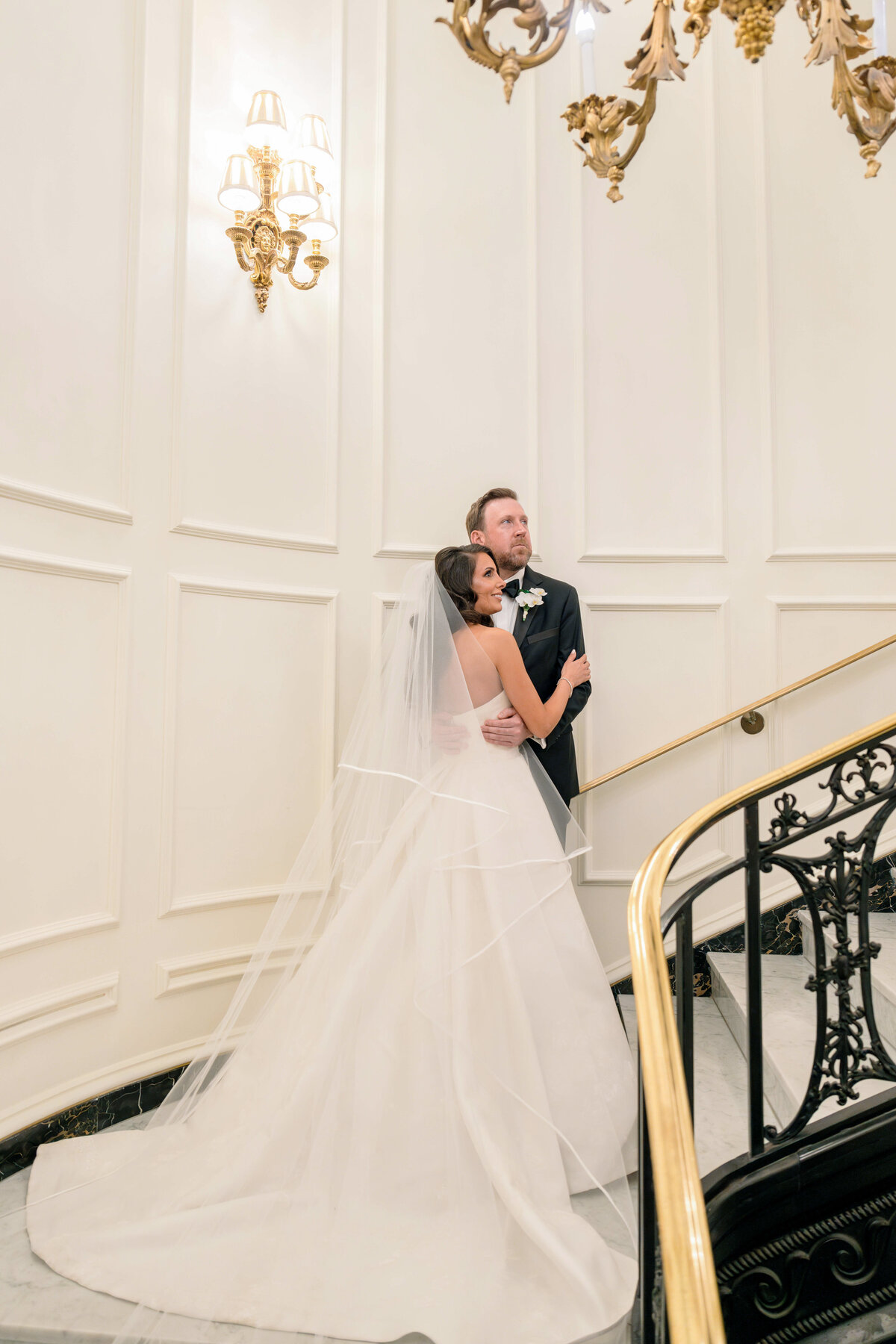A bride in a flowing gown and veil embraces a groom in a dark suit as they stand on an ornate staircase with elegant gold and black railings. A decorative light fixture and paneled walls add to the sophistication of the setting.