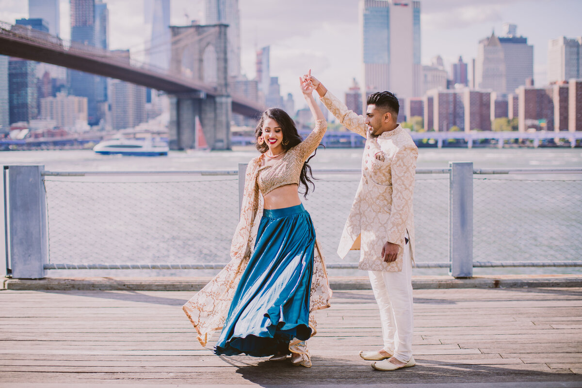 An engaged couple twirl near the carousel at brooklyn bridge park dumbo with the bridge in the background