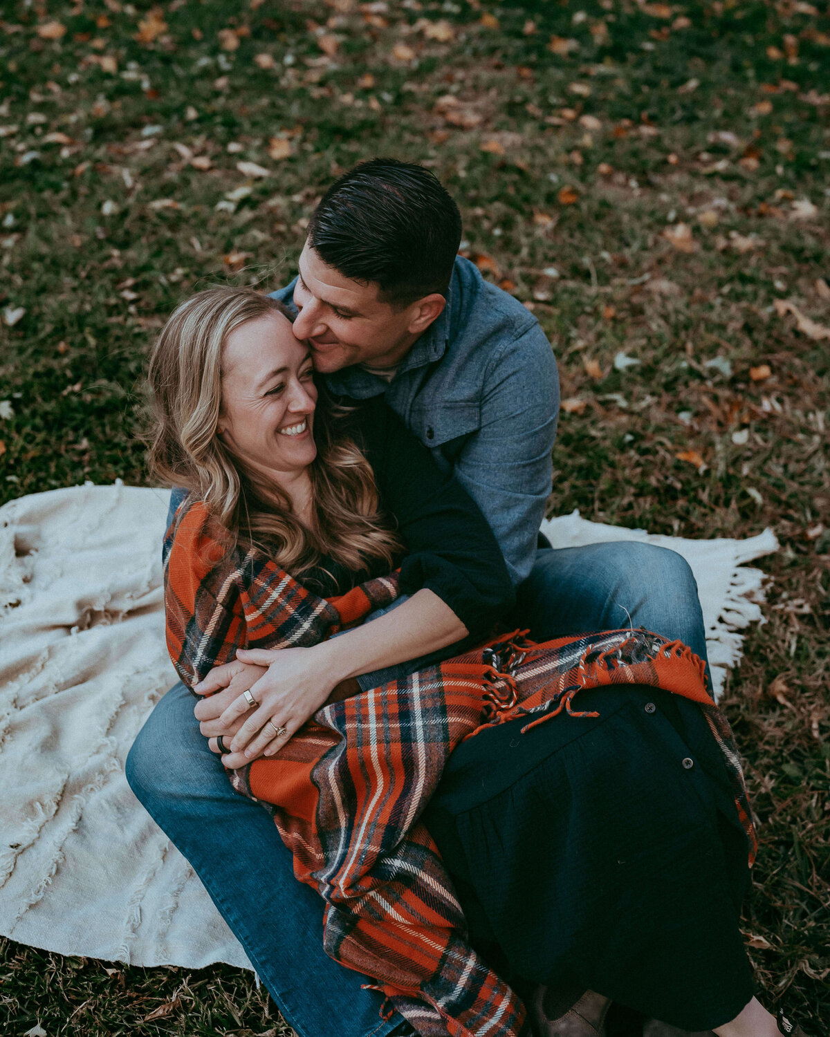 A husband and wife playfully sit on a white blanket on their family farm as they watch their three boys run around just being kids.