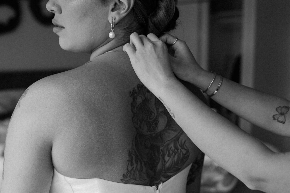 Black and white image of a woman in a strapless wedding gown being helped with her neckalce, showcasing a detailed tattoo on her back