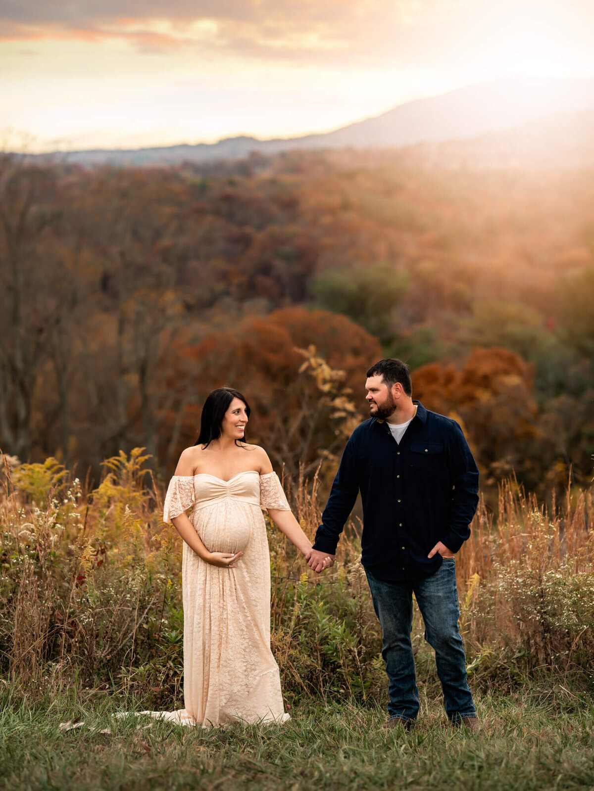 An expecting couple hold hands and look at each other while standing in a field with gold grass