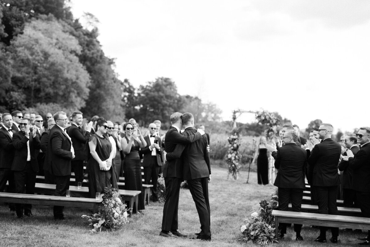 Two men in tuxedoes lean into one another