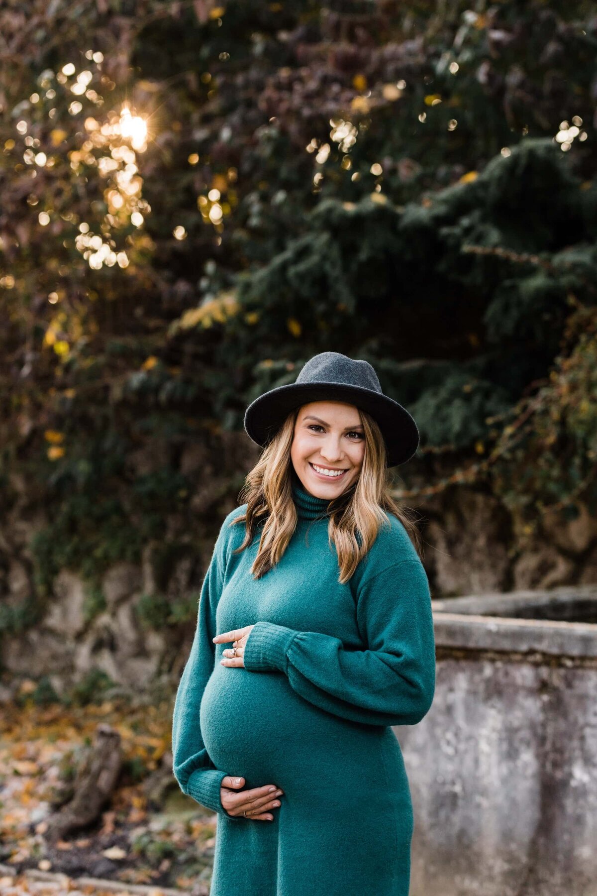 A smiling pregnant woman in a green dress and a gray hat, captured by a Pittsburgh maternity photographer, holding her belly outdoors.
