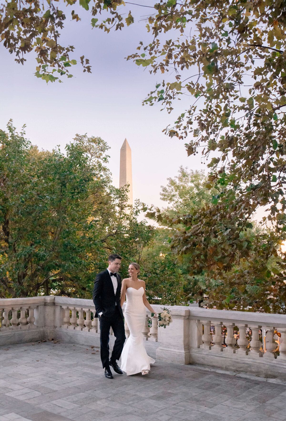 A couple in elegant attire walks hand in hand on a stone terrace surrounded by trees. The woman wears a white wedding dress, and the man is in a black suit. The Washington Monument is visible in the background.