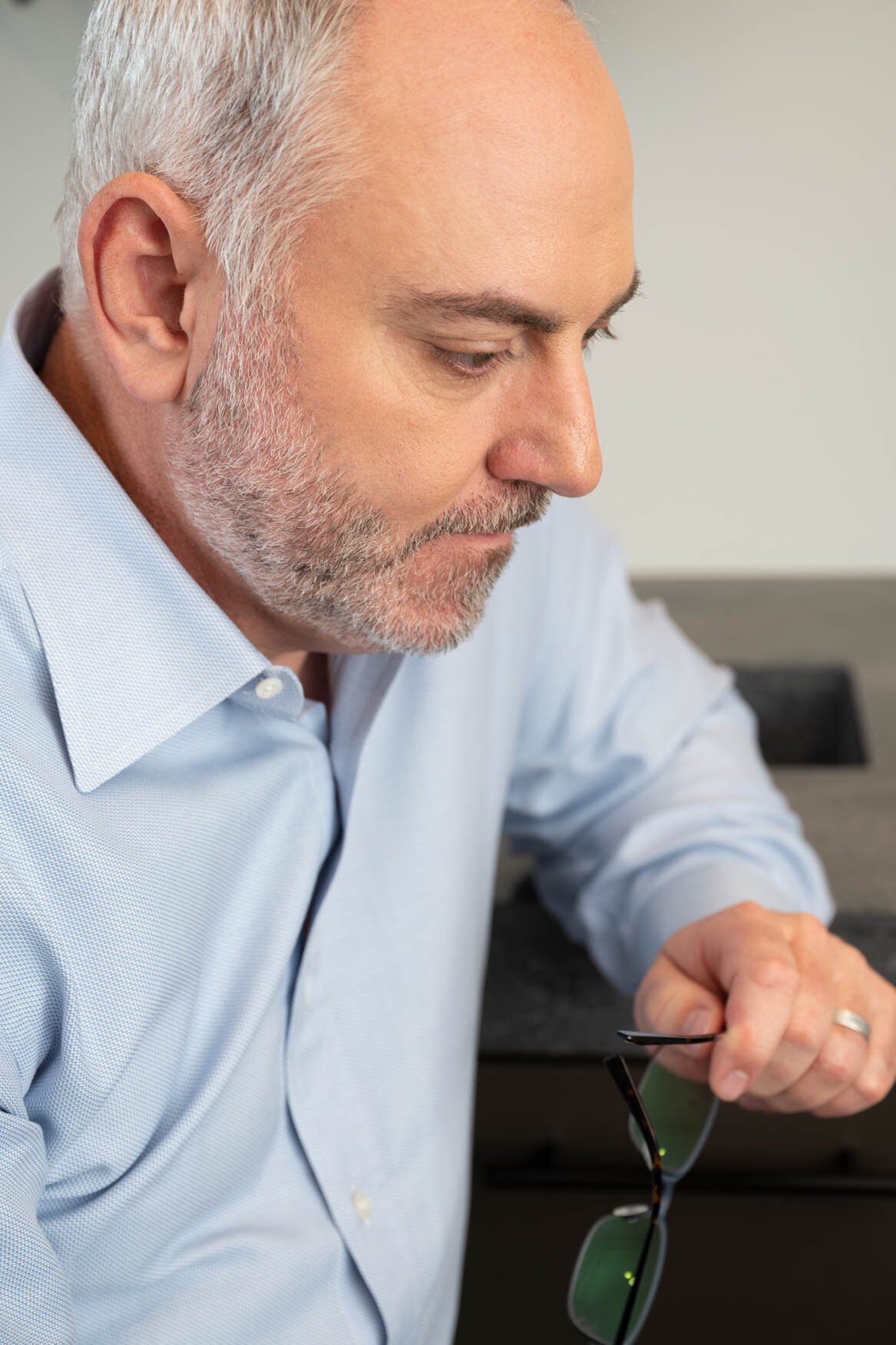 Close-up portrait of a man in a light blue dress shirt holding glasses, captured in a moment of focus and reflection with a neutral background