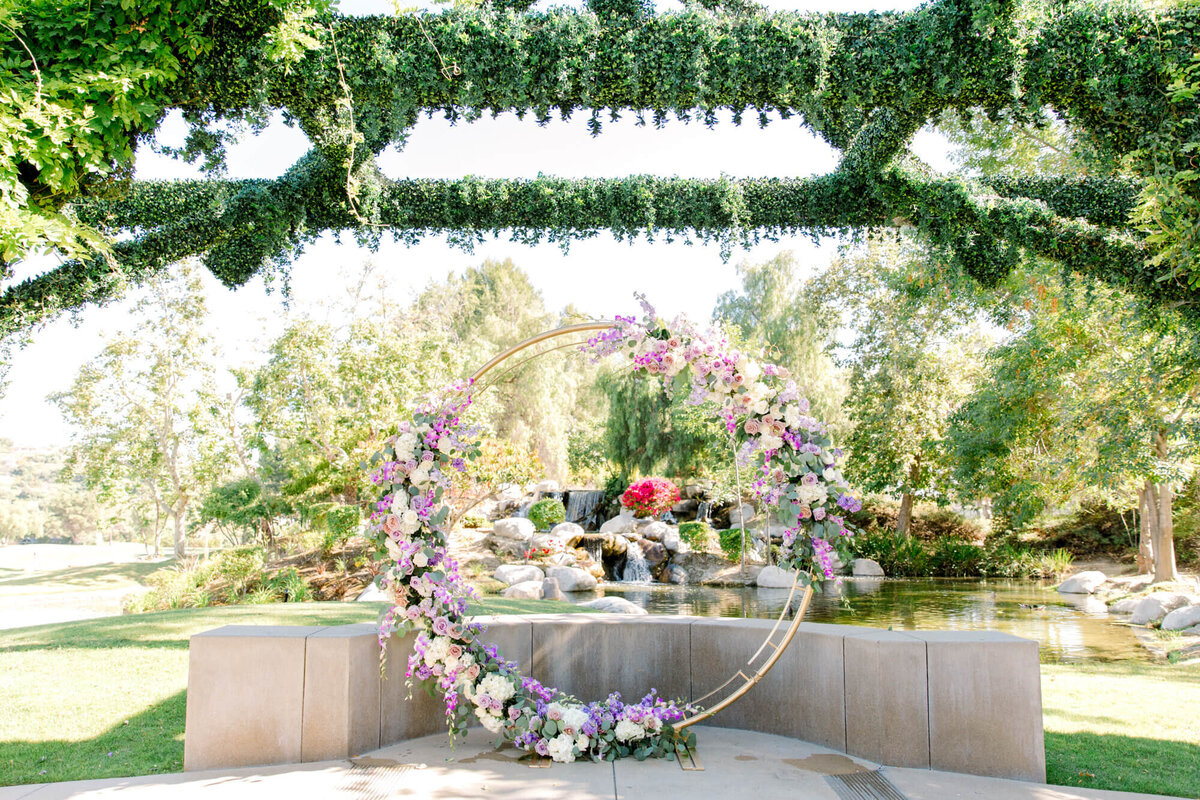 A circular floral arch with pink, purple, and white flowers stands in a garden, framed by lush greenery and a backdrop of a serene pond and waterfall.