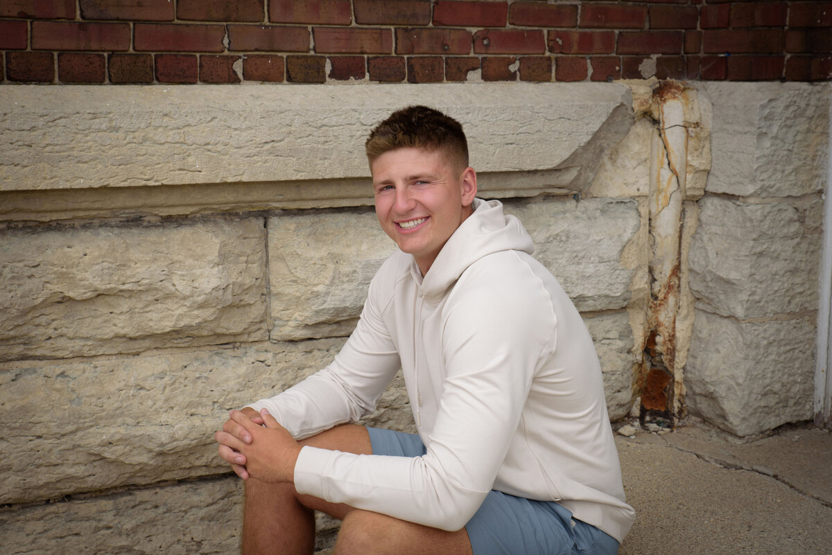 De Pere High School senior boy wearing blue shorts and a cream hooded sweatshirt sitting in an urban setting at The Depot in Downtown Green Bay, Wisconsin