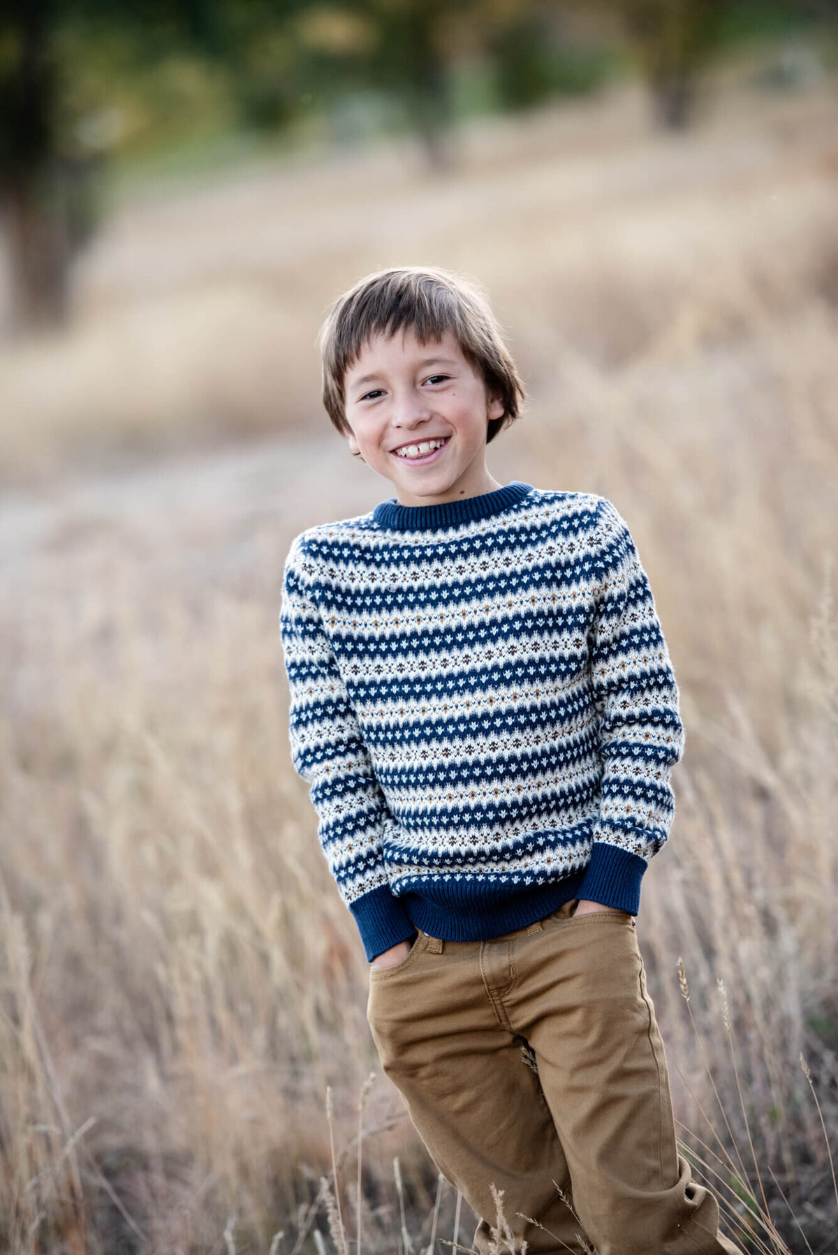 A young boy in a stripe sweater and khakis stands smiling in a field at sunset with hands in pockets