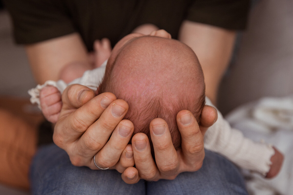 Livsstilsfoto av far som holder babyen sin på knærne sine, hodet ligger i hendene hans. Fotografert slik at man ser hodet til babyen mot kamera.