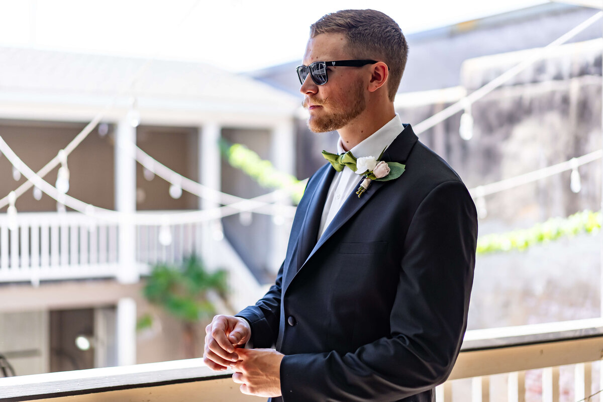 A man in a black suit, white shirt, and sunglasses stands on a balcony decorated with string lights, wearing a boutonnière and looking thoughtfully into the distance.