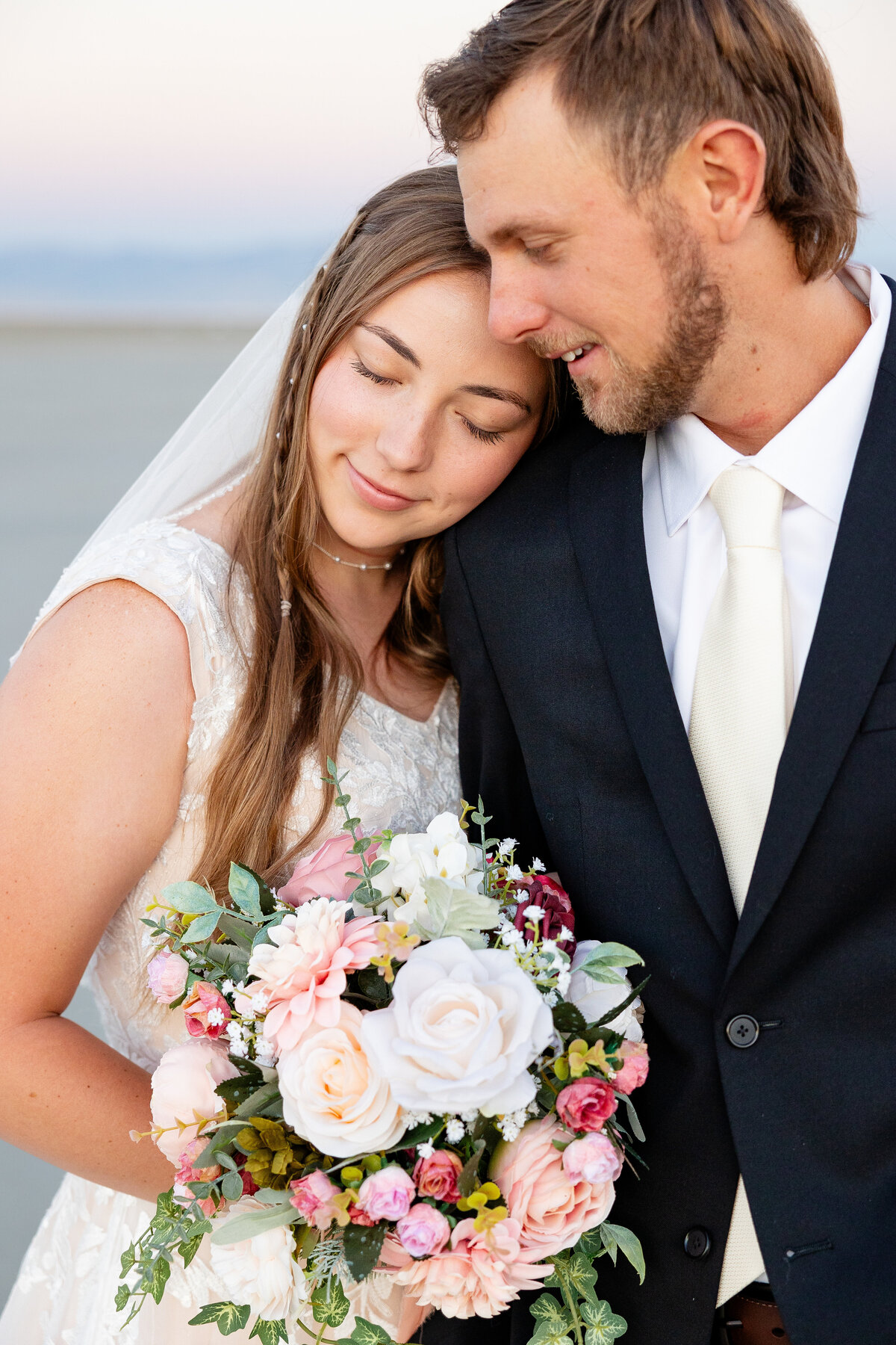 Bride leans her head on the groom's shoulder, holding a bouquet of pastel flowers.