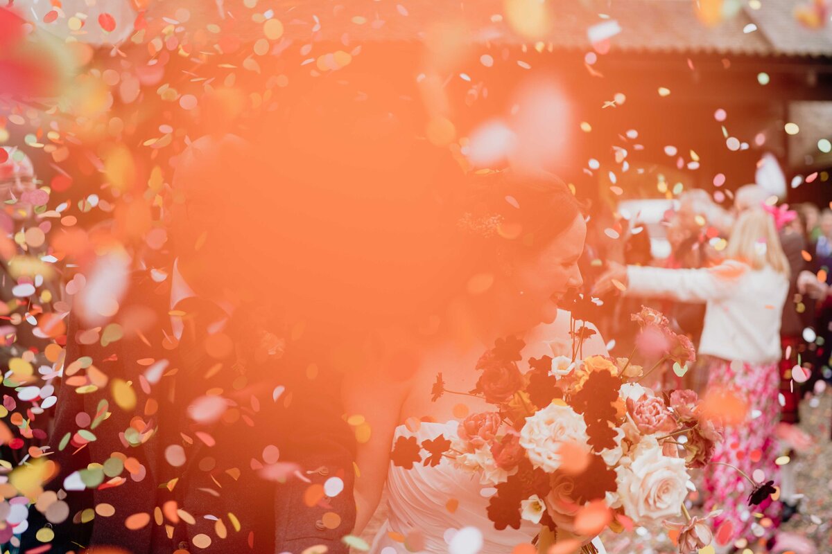 A bride in a white dress smiles, bouquet in hand, as colourful confetti falls directly in front of the photographer’s camera lens. The photo is blurred in bright orange and the festive outdoor celebration is filled with joy and laughter from friends and family celebrating in the background. The courtyard of Charleton Fruit Farm in Aberdeen is see all around them.