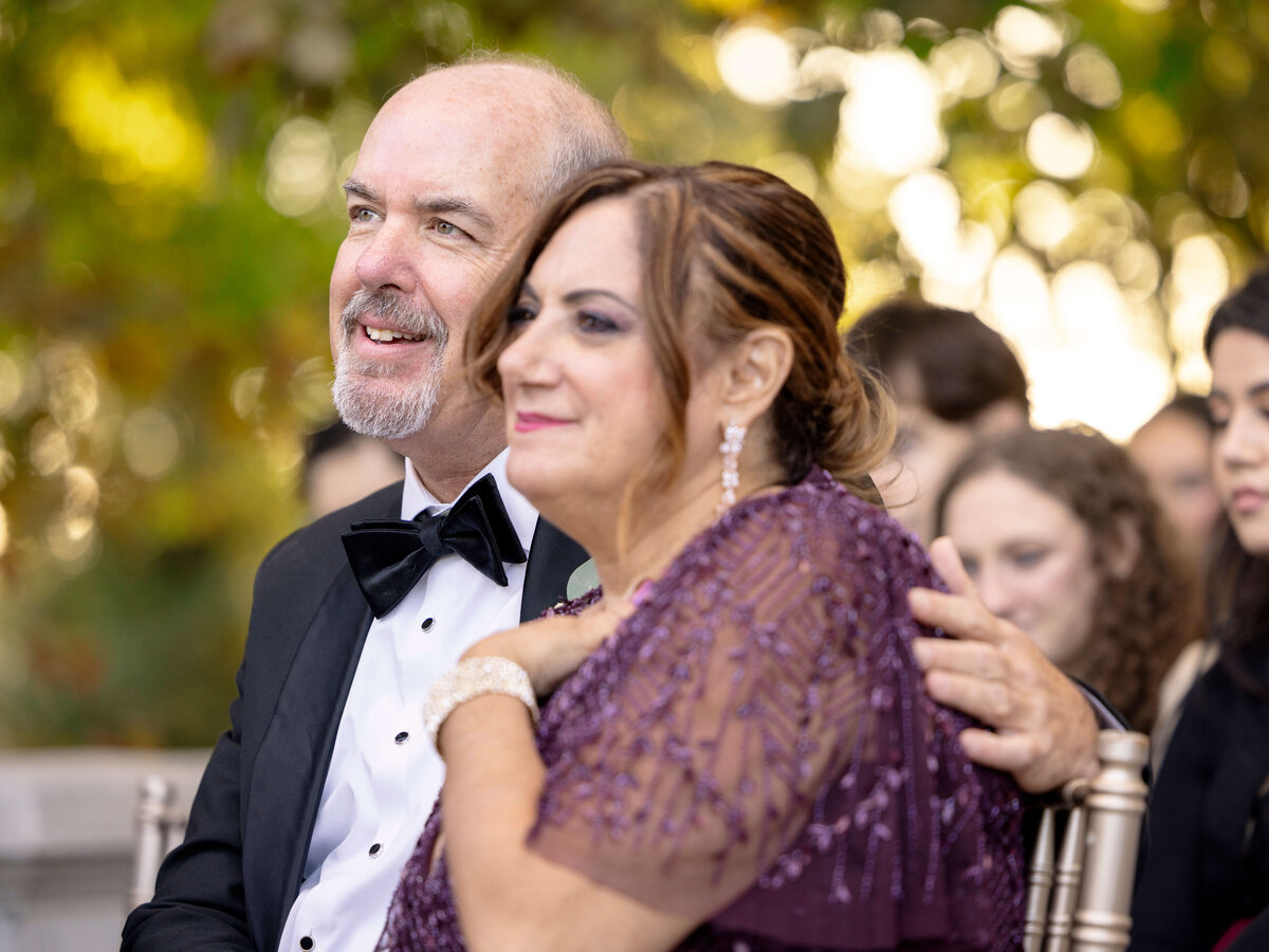 A smiling couple, dressed elegantly, sits together outdoors. The man wears a tuxedo, and the woman is in a purple dress with beadwork. They are surrounded by other people in a wooded setting, enjoying what seems to be a celebratory event.