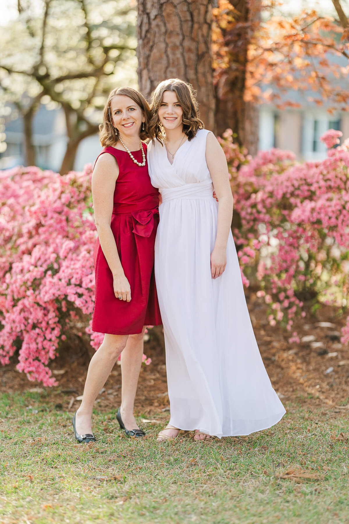 Two women, captured by a Fayetteville NC family photographer, stand outdoors in front of a tree and pink flowering bushes. One wears a red sleeveless dress and the other a white sleeveless gown. Both are smiling with their arms around each other, set against a backdrop of a house and trees.