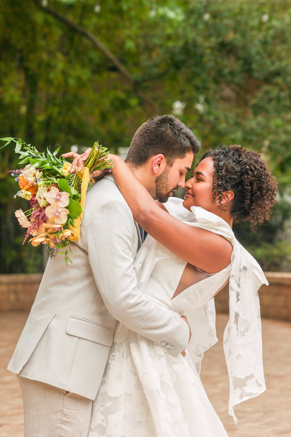 bride wearing white dress with bows on sleeves while holding colorful bouquet with arms around groom wearing tan suit