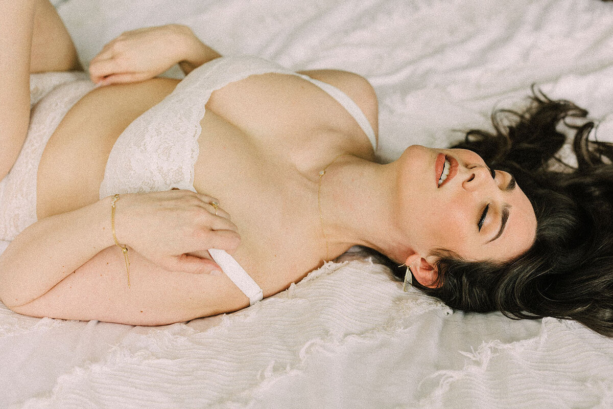 A woman poses on a bed for a bridal boudoir photoshoot while wearing white lingerie.