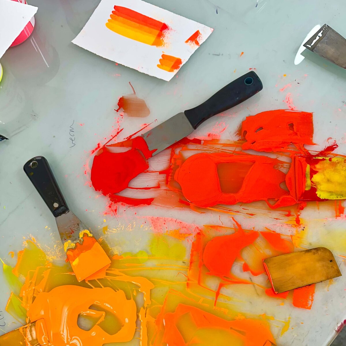 A messy mixing palette of orange, yellow, and red etching inks with palette knives and swatch tests, capturing the vibrant, hands-on energy of pigment mixing.