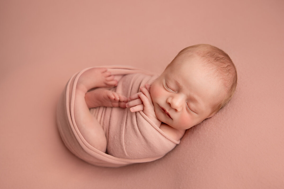 Newborn baby girl wrapped in a light pink wrap with her hands and feet exposed on a matching backdrop.
