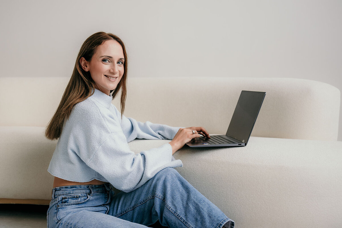 Woman sitting on the ground while typing on a laptop propped up on a sofa.