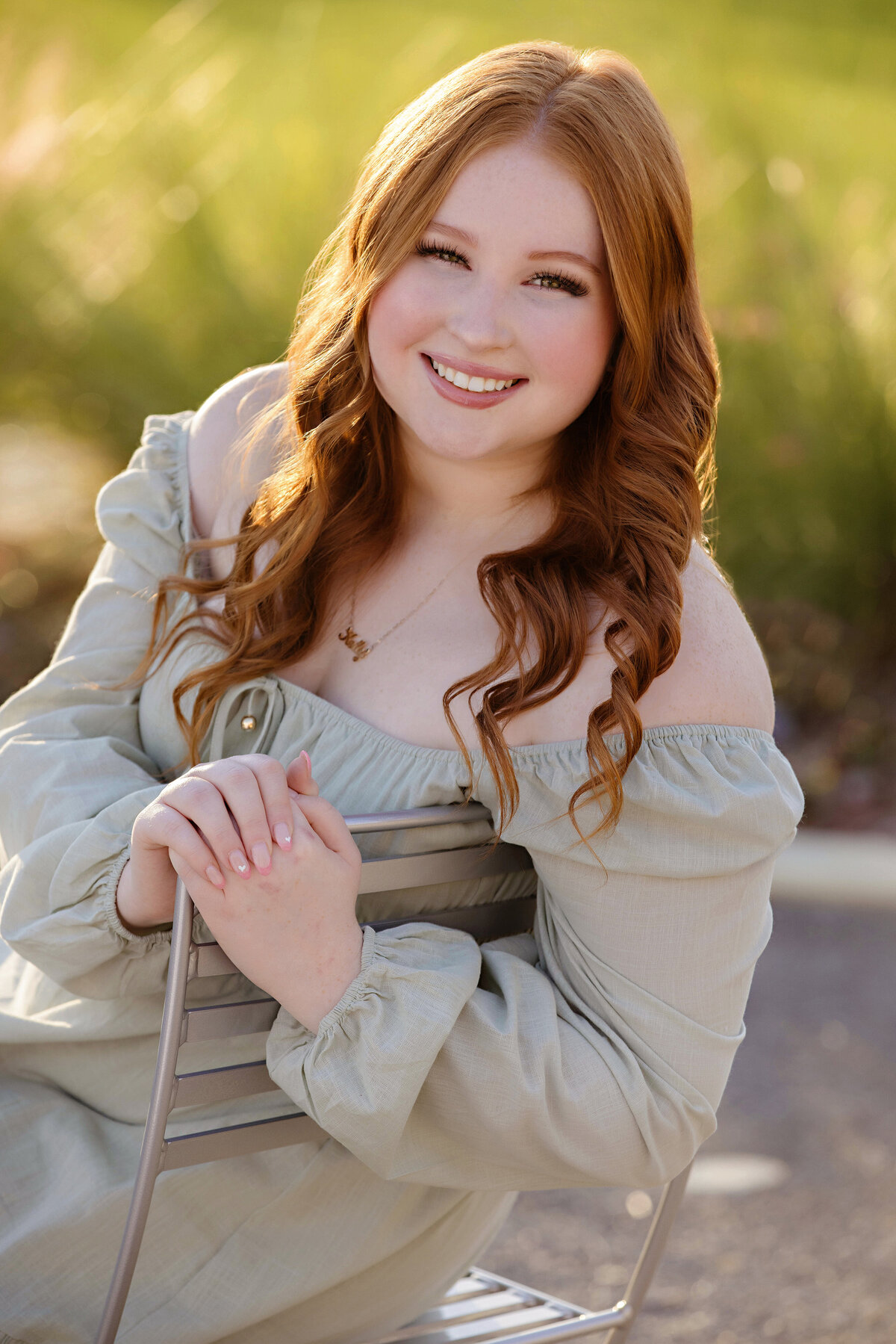 High school senior in sage green dress seated in metal chair with tall grass behind her and sunflare glowing through strands of hair