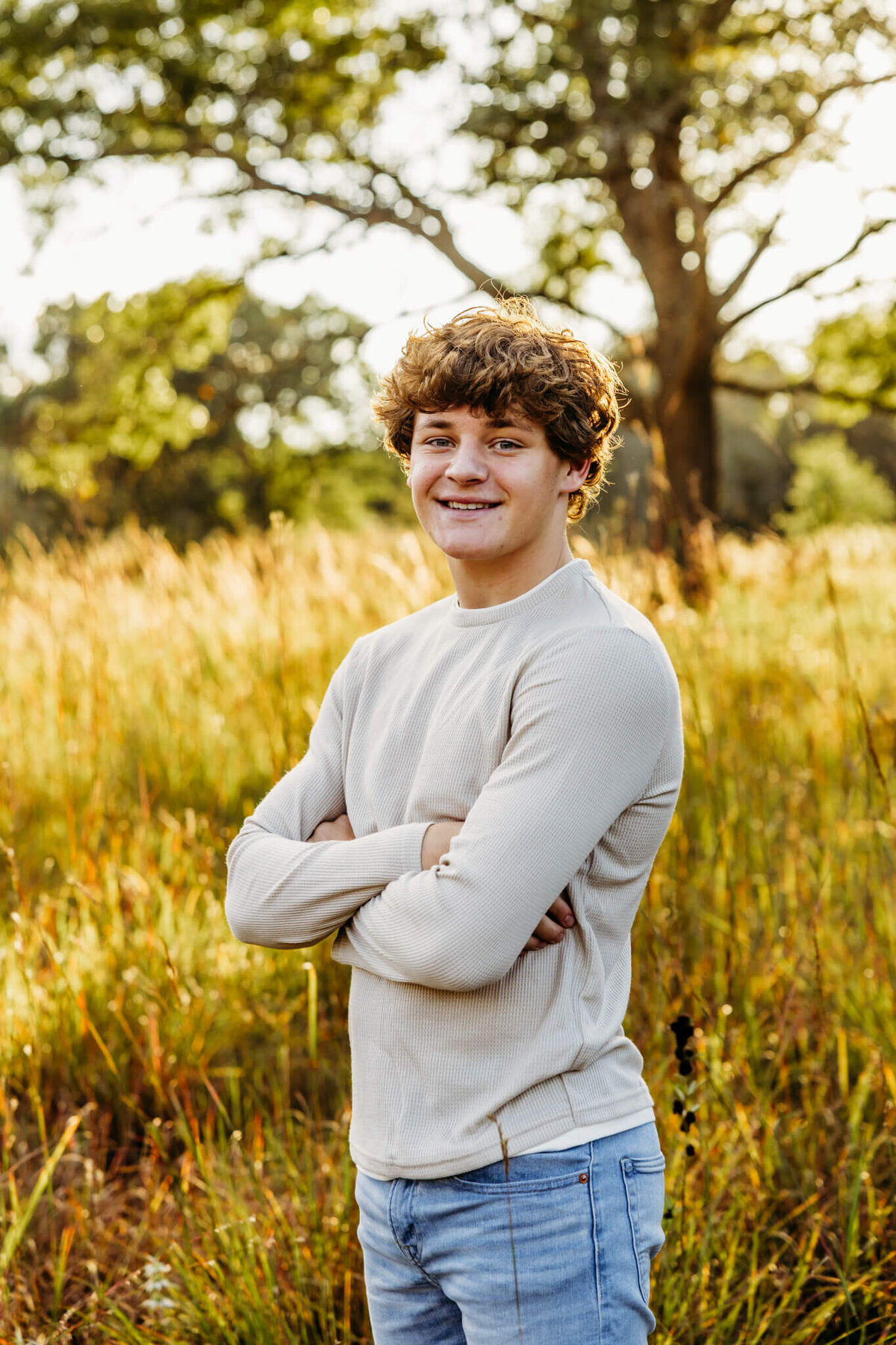 young man in a white long sleeve top crossing his arms and smiling as he stands in a field at sunset.