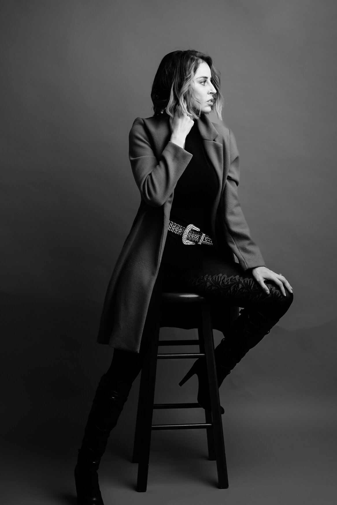 a black and white self-portrait of a woman sitting on a stool wearing a black outfit and a dark long coat