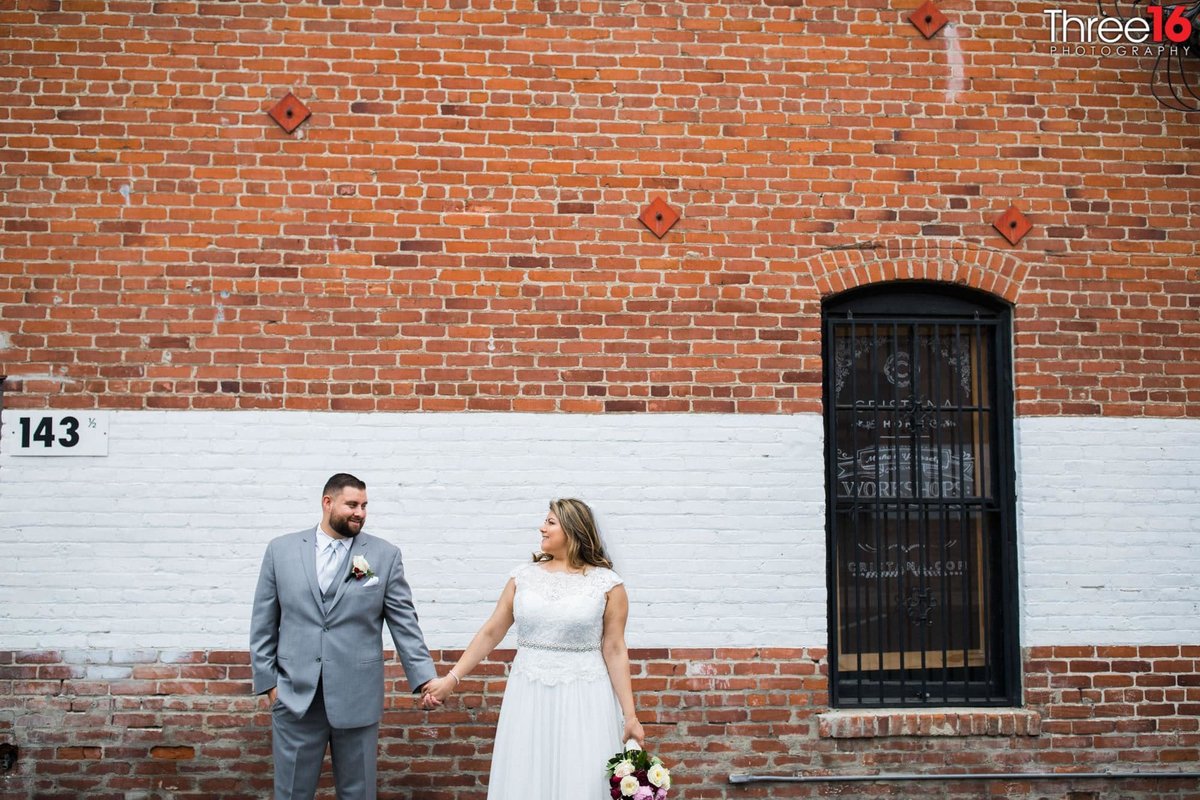 Bride and Groom hold hands as they pose against a brick wall in a back alley in Downtown Orange