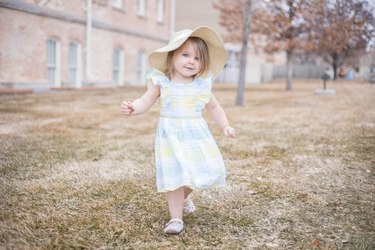 Two year old toddler girl in a bllue and yellow dress and a sunhat running on a grassy lawn