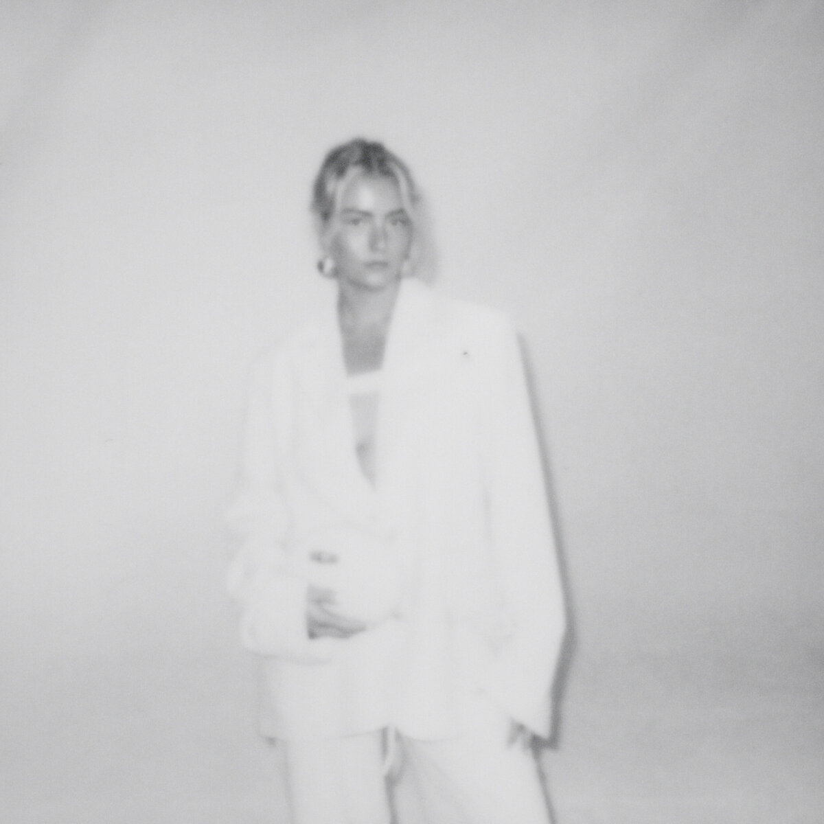 A model standing against a plain white backdrop wearing a white blazer, hoop earrings and holding a white clutch.