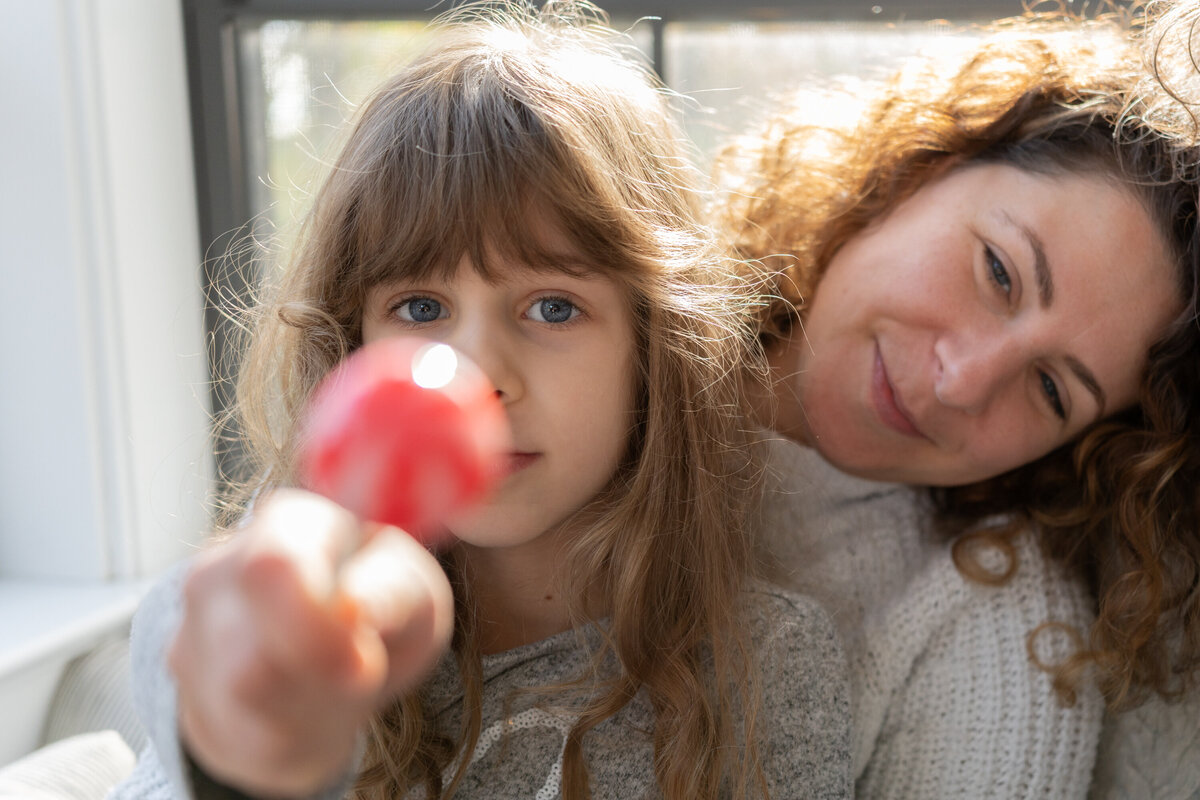 little girl holds lolipop towards camera while mom looks lovingly over her shoulder